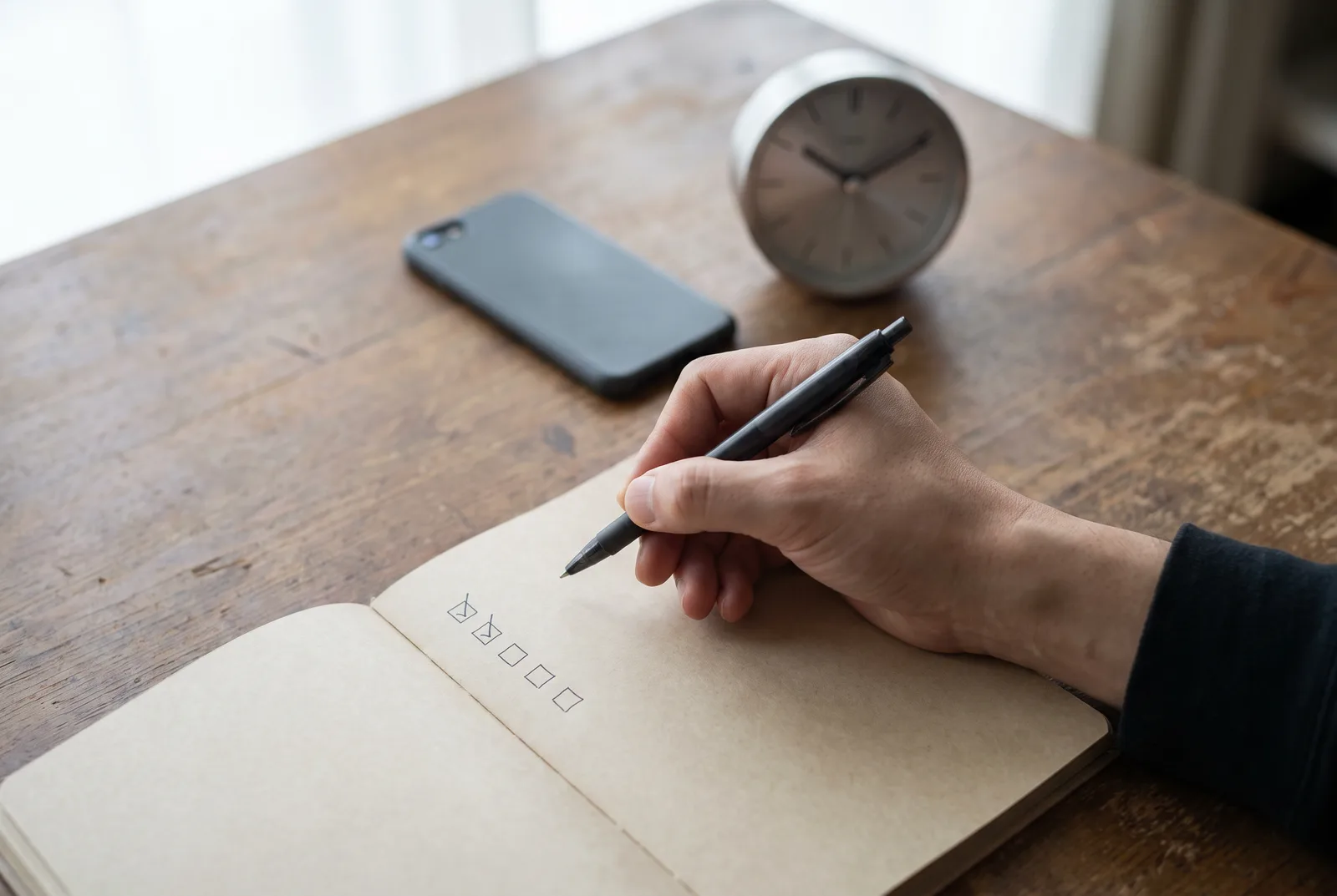 A hand holding a pen over a notebook page with a column of small boxes, two of them ticked, beside a phone lying face down and a small desk clock.