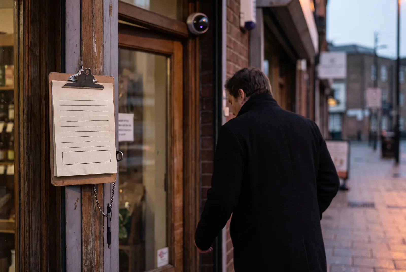 A shop entrance at dusk. A clipboard with a completely blank consent form and an unused pen hangs by the door, a dome camera glows faintly above it, and a shopper walks in without looking at either.