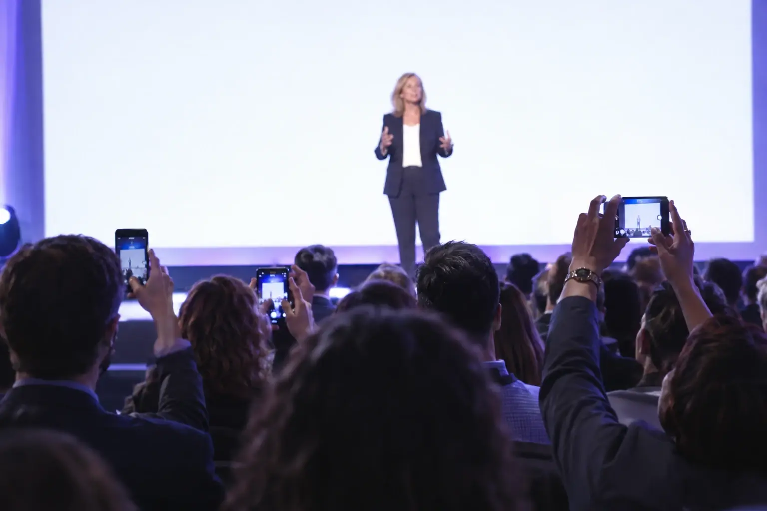 A woman in her late forties speaks on a conference stage under bright lights while audience members in the foreground hold up their phones to photograph her.
