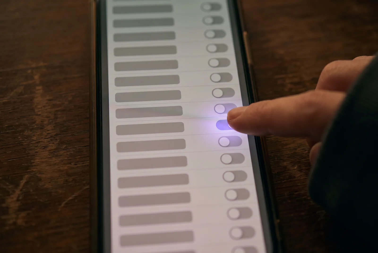 Close-up of a phone showing a very long list of grey toggle switches. Deep in the list, one switch is on and glowing violet, with a thumb hovering over it.