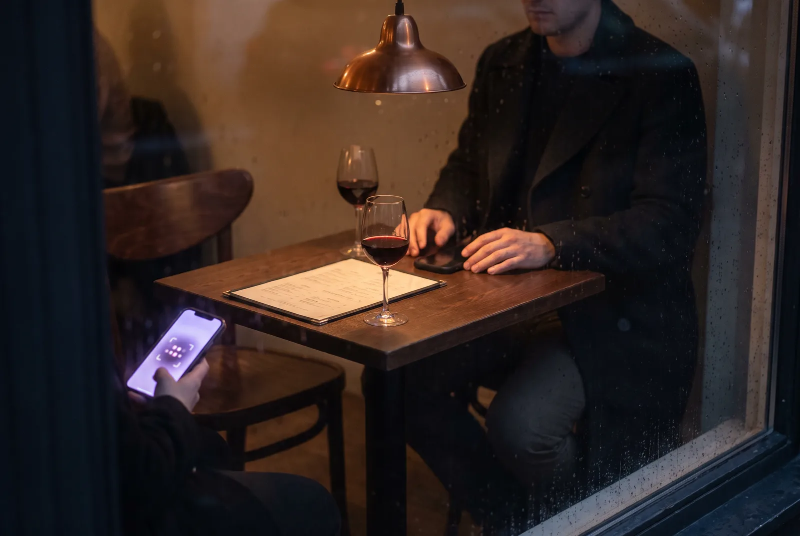 A café table for two at evening. One person is seated across the table, seen only from the shoulders down. On the near side, an empty chair and a phone lit with a soft violet face-match overlay.